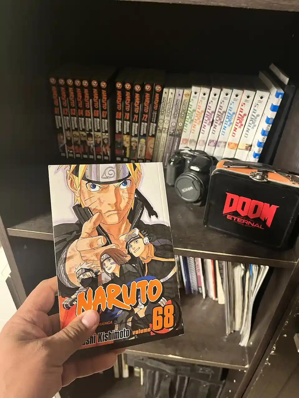 A hand holds up the Naruto manga volume 68, with a bookshelf in the background containing many other Naruto volumes, a Nikon…