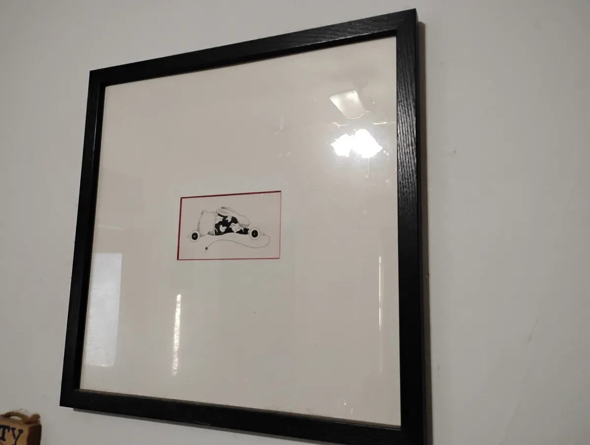 A framed picture on a wall shows a simple black-and-white drawing of a rabbit inside a car with wheels, enclosed in a red…