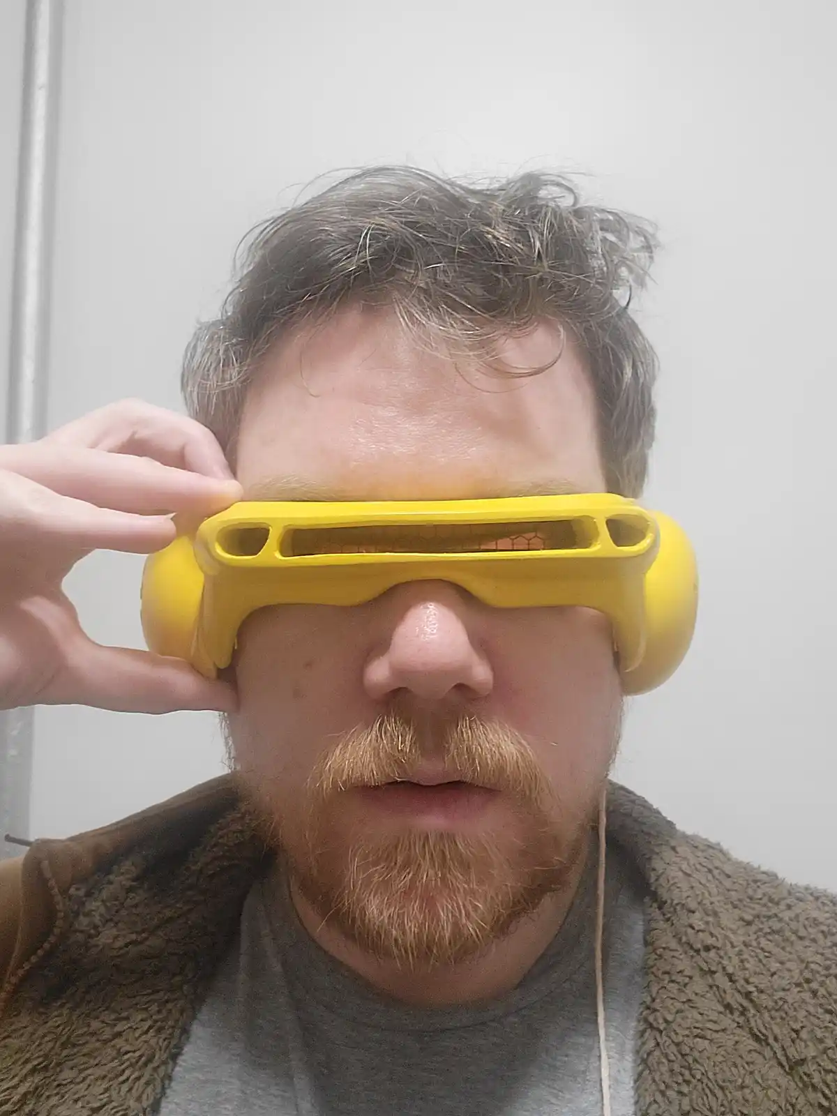 A man with a reddish beard and grayish hair holds a bright yellow, bulky, futuristic-looking pair of glasses over his eyes…