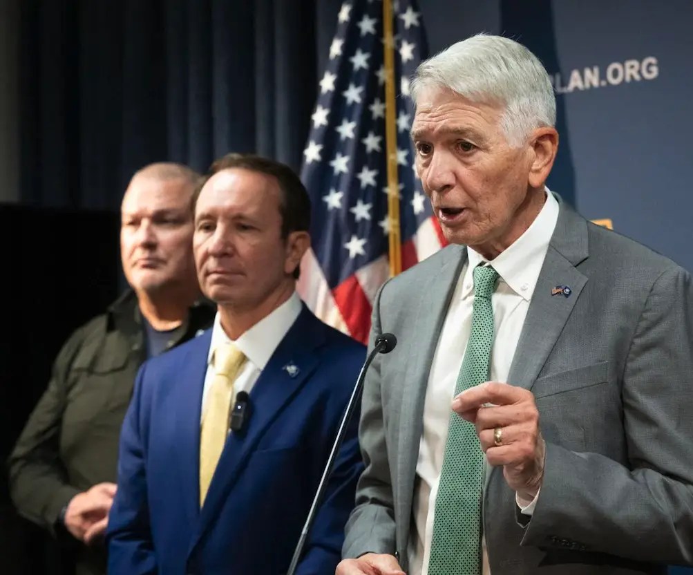 Three men stand at a press conference with an American flag and a blue backdrop featuring 'PLAN.ORG'. The man in the…