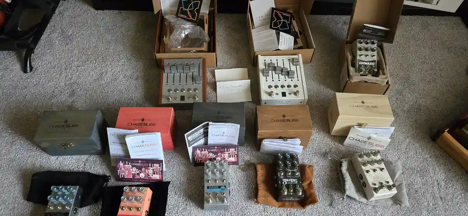 An overhead shot displays multiple Chase Bliss Audio guitar pedals and their packaging spread across a carpeted floor…