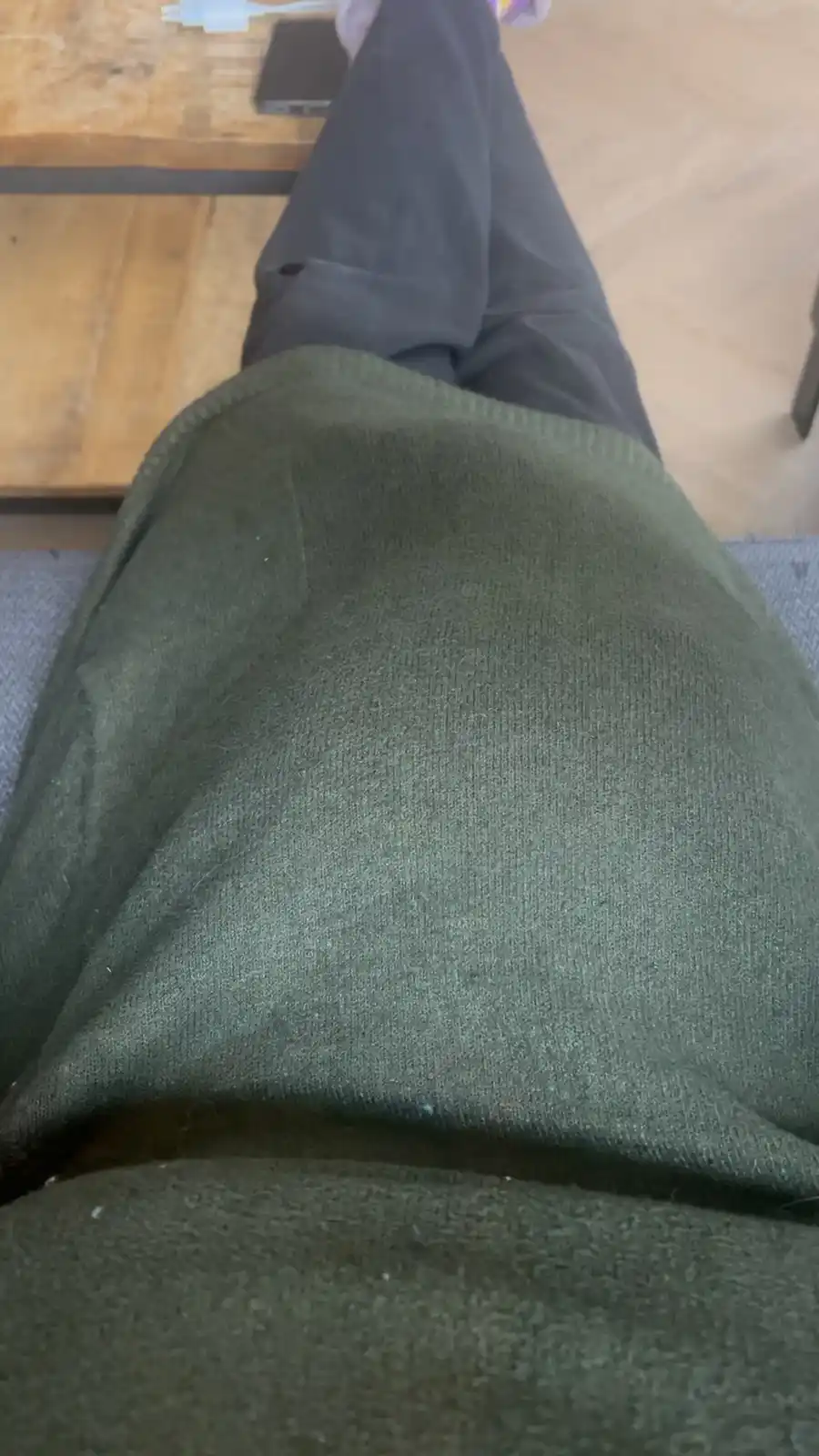 A first-person view of someone sitting on a gray couch, wearing a dark green sweater and dark pants, with a wooden table and…
