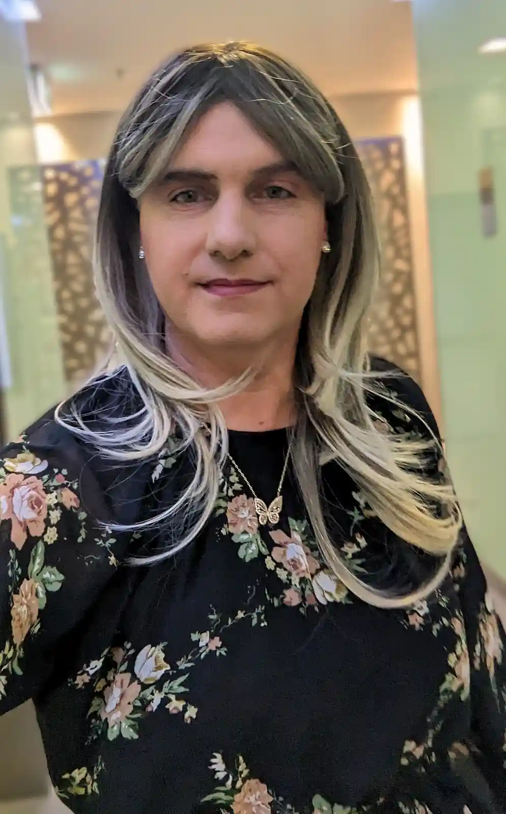 A person with shoulder-length, layered hair featuring dark roots and blonde highlights, wearing a black floral top and a…
