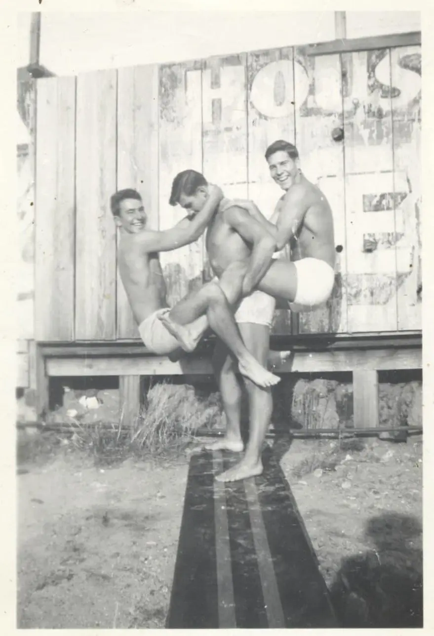 A black-and-white vintage photo shows three shirtless men in swim trunks playfully posing on a wooden platform. One man is…