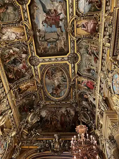 The image shows an ornate, gilded ceiling filled with numerous classical paintings depicting mythological scenes, framed by…
