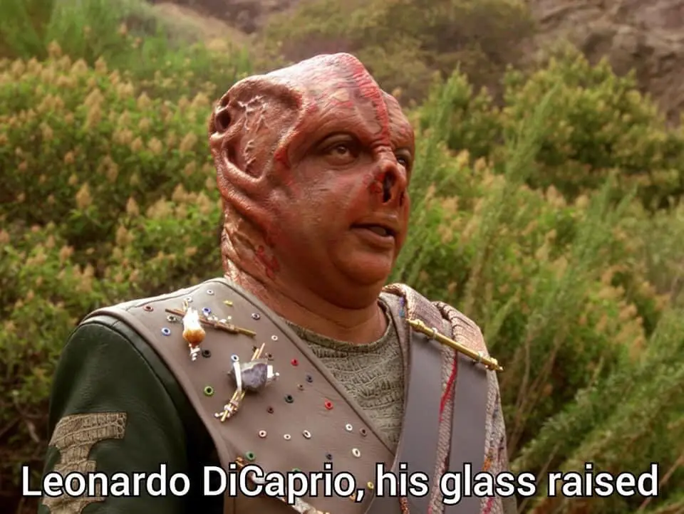 A close-up of a character with a red, textured alien-like face and a decorated vest, standing in a bushy outdoor setting…