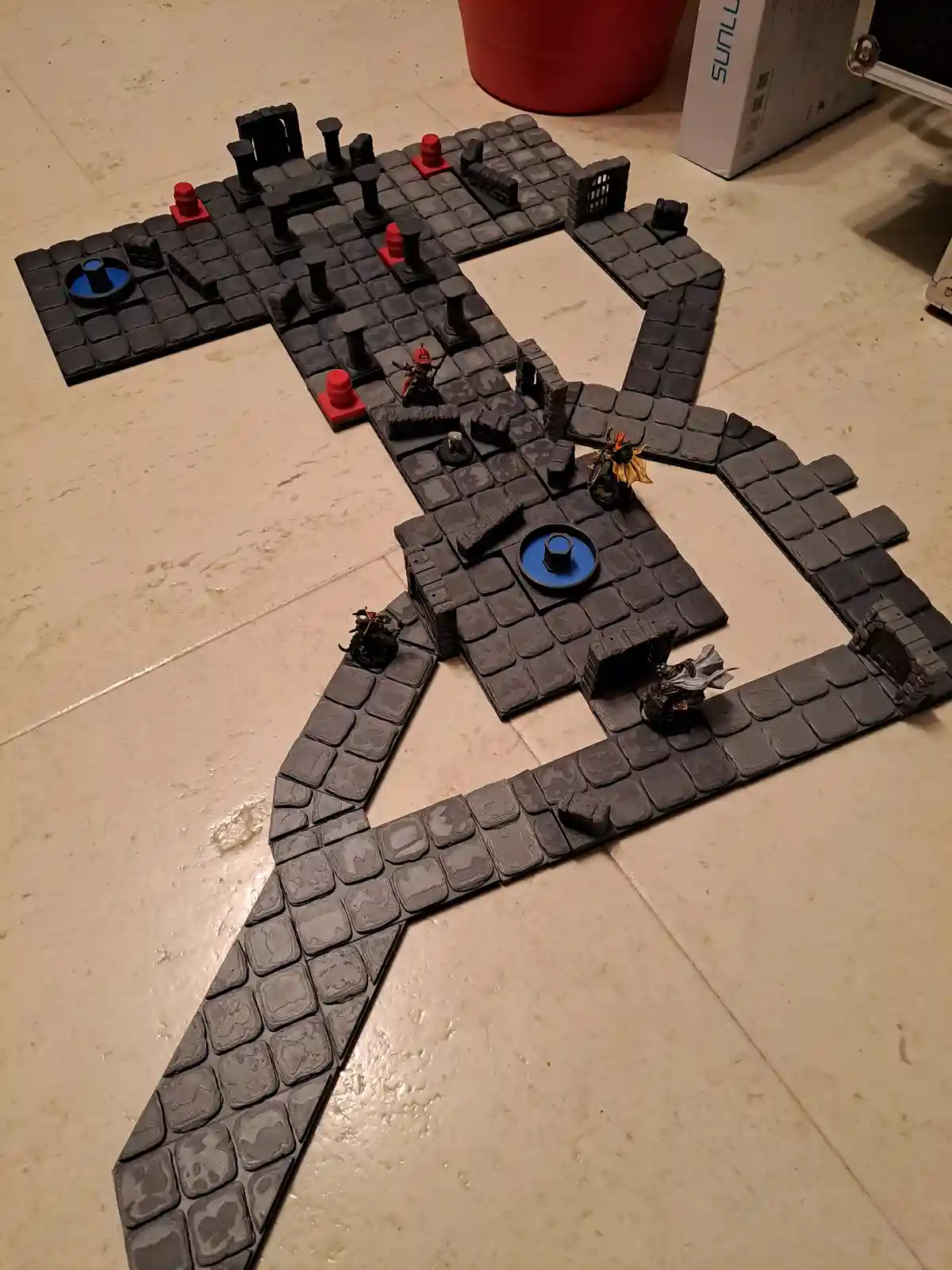 A tabletop miniature game setup on a tiled floor, where black stone tiles are arranged to form the shape of the letter 'X'…