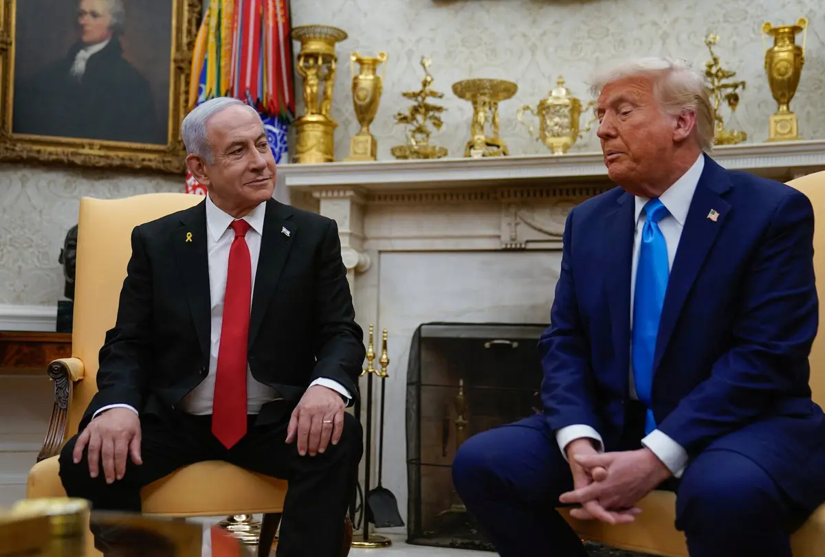 two men in suits sit facing each other in an ornate room with a fireplace and golden trophies behind them. The man on the…