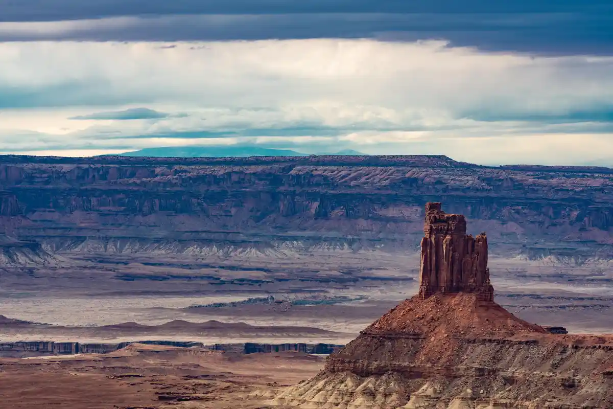 A dramatic desert landscape featuring a prominent red rock butte in the foreground, with layered canyon walls and distant…