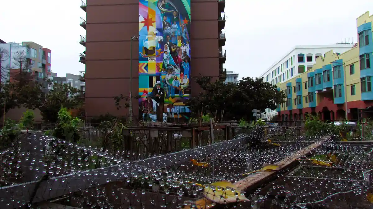 A rainy urban scene viewed through a window with water droplets, showing colorful buildings and a large mural on a brown…