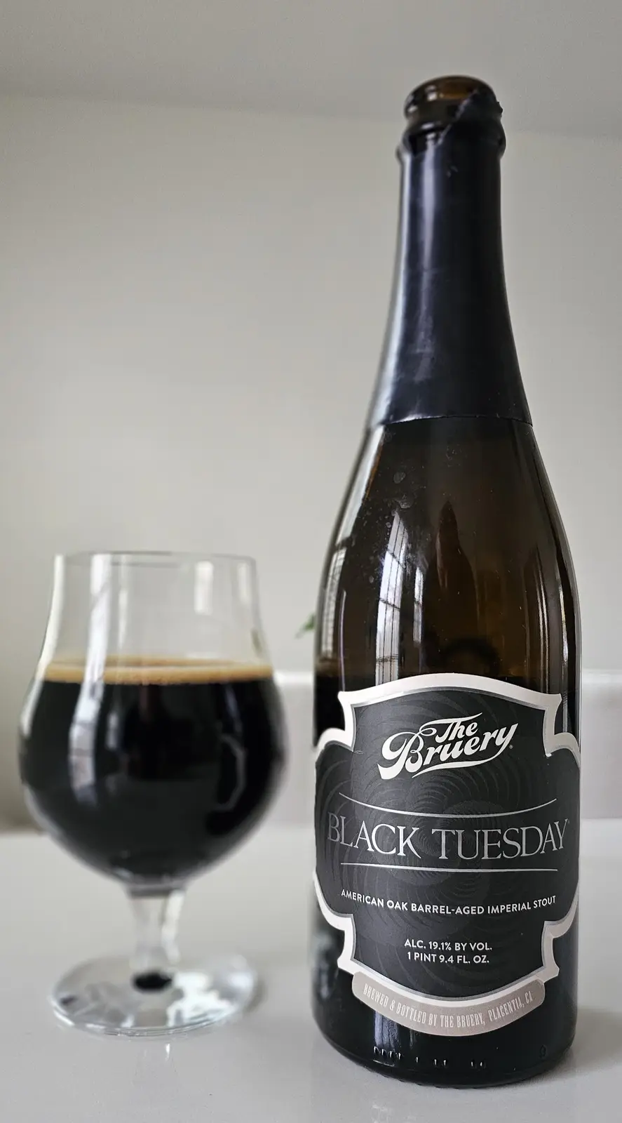 A dark brown bottle of 'The Bruery' beer labeled 'BLACK TUESDAY' sits next to a glass filled with the same dark beer, both…