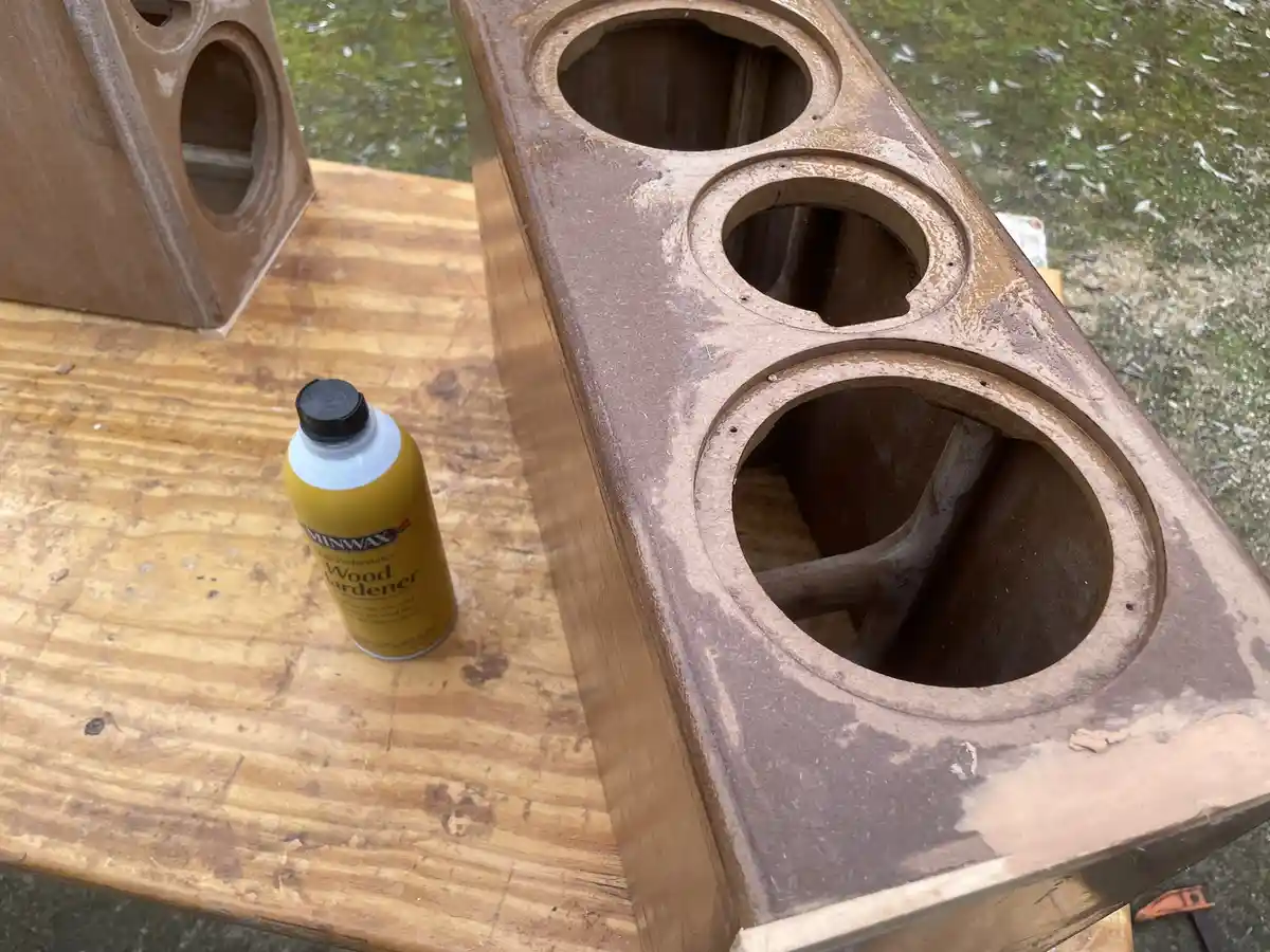 A yellow can of Minwax Wood Hardener sits on a wooden workbench next to a large, unfinished wooden box with three circular…