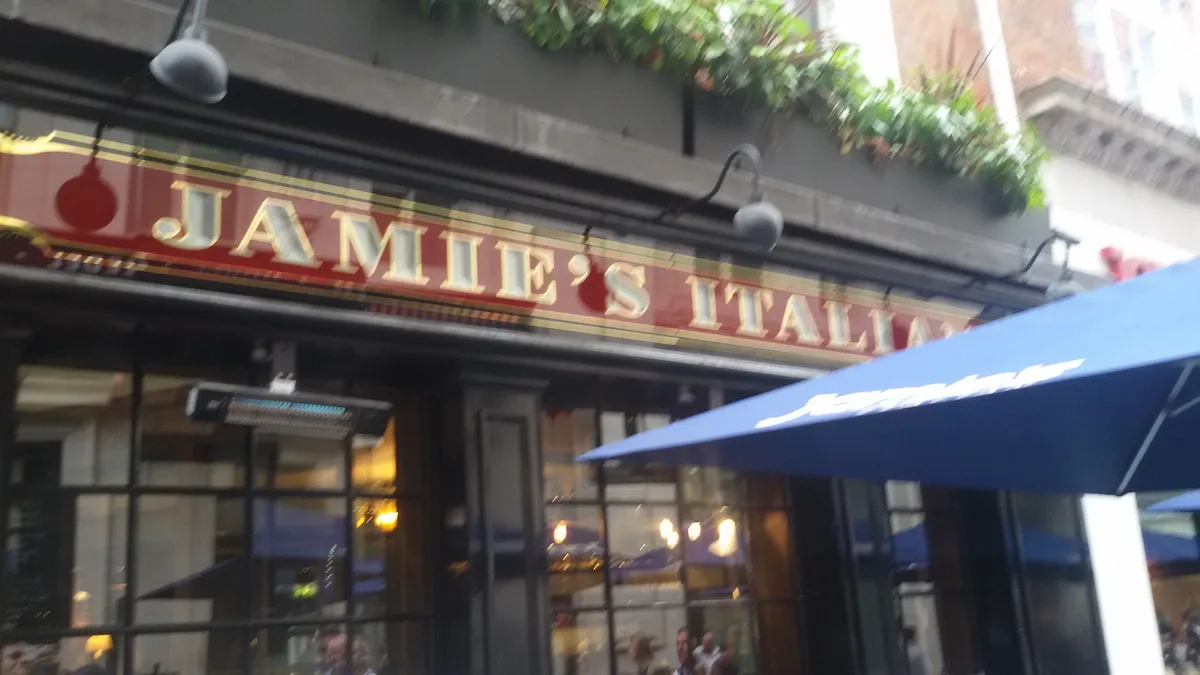 A blurry photo of a restaurant's exterior sign that reads 'JAMIE'S ITALIAN' in gold letters on a red background, with a blue…