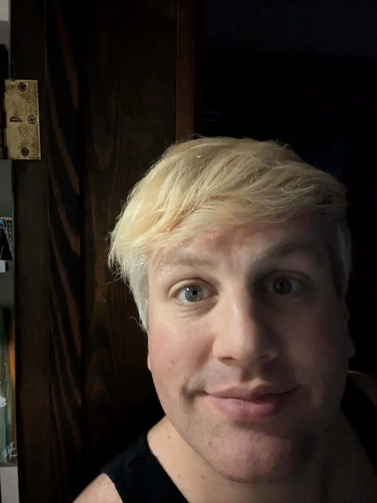 A close-up selfie of a man with bright blonde hair, looking directly at the camera with wide eyes and a slight smirk…