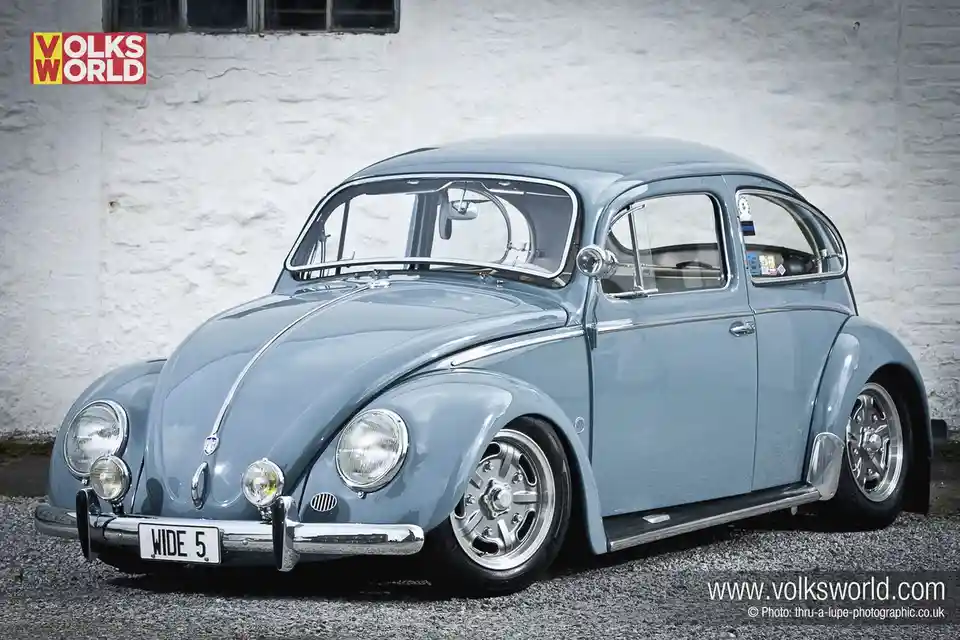 A light blue classic Volkswagen Beetle is parked on gravel in front of a white stucco wall. The car has chrome trim, custom…