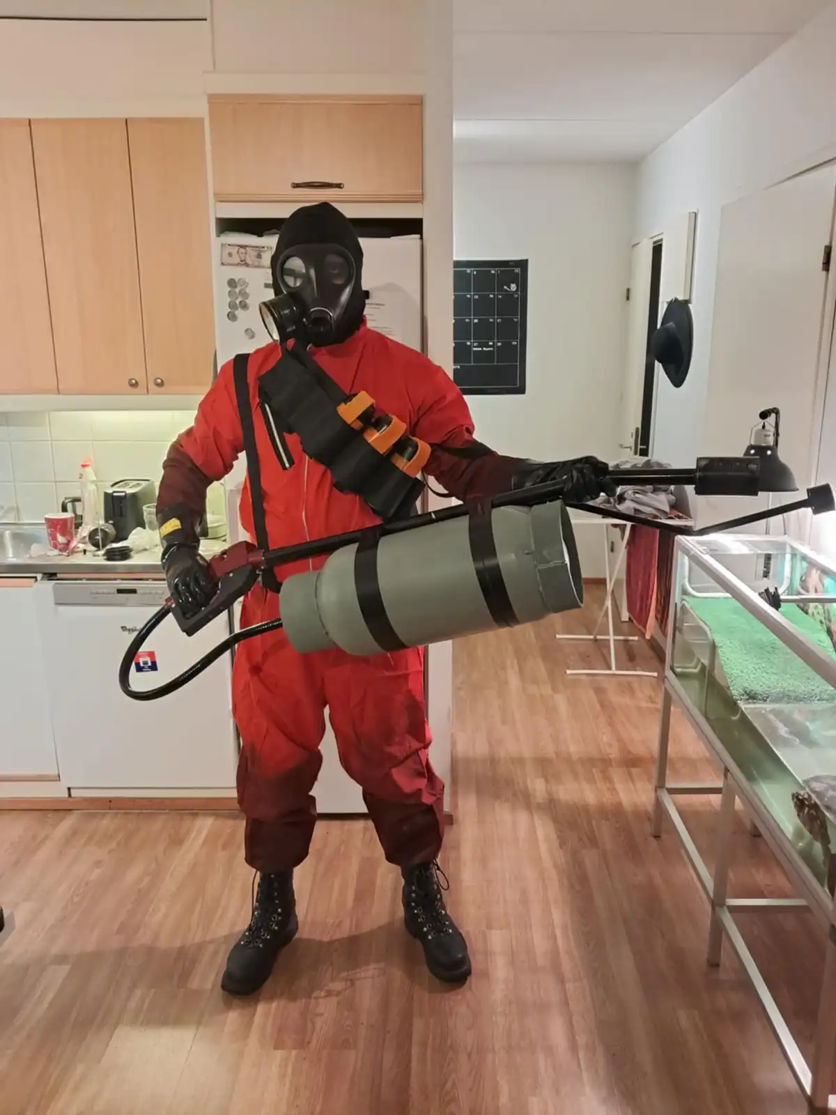 A person in a red hazmat suit, black gas mask, and black gloves stands in a kitchen holding a large, homemade-looking…