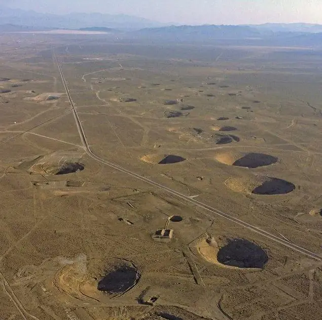 an aerial photograph shows a long straight road cutting through a vast, arid desert landscape dotted with numerous large…