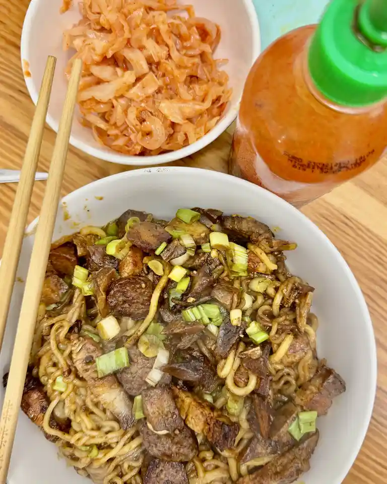 A close-up photo shows a bowl of stir-fried noodles with meat and chopped green onions, next to a bowl of kimchi and a…