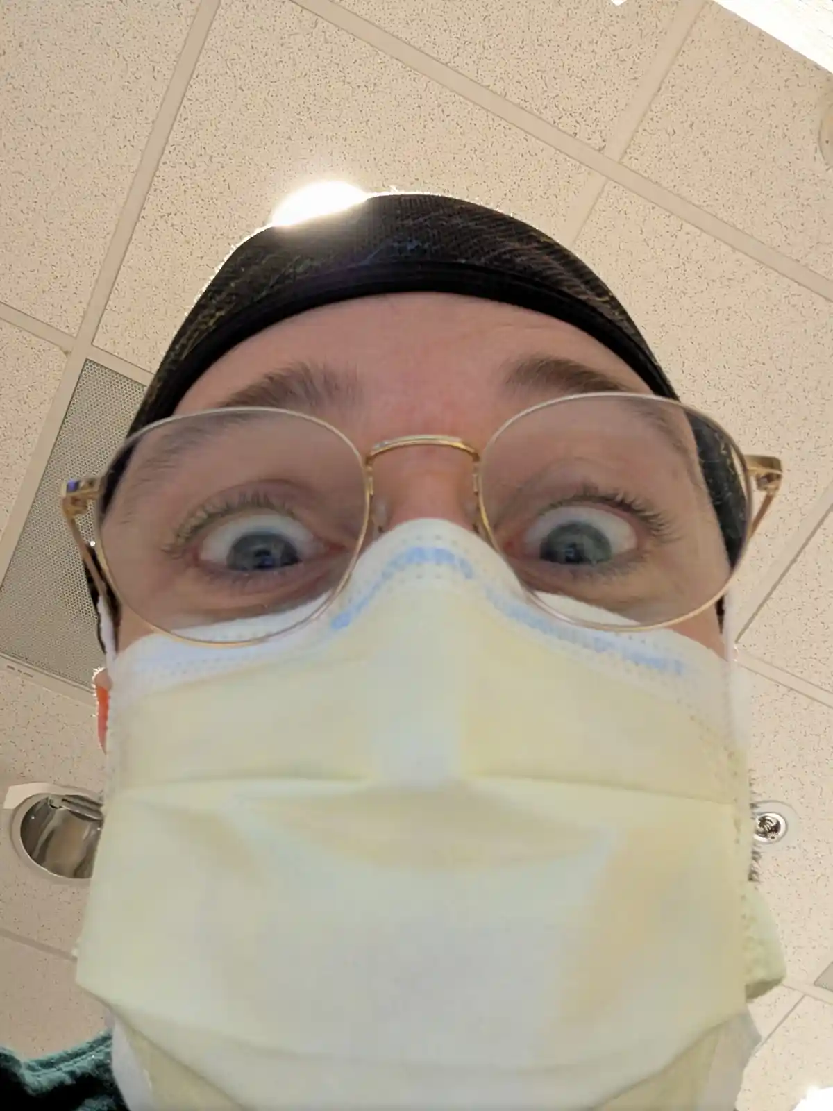 A person wearing a black head covering, gold-rimmed glasses, and a light-colored surgical mask looks directly at the camera…