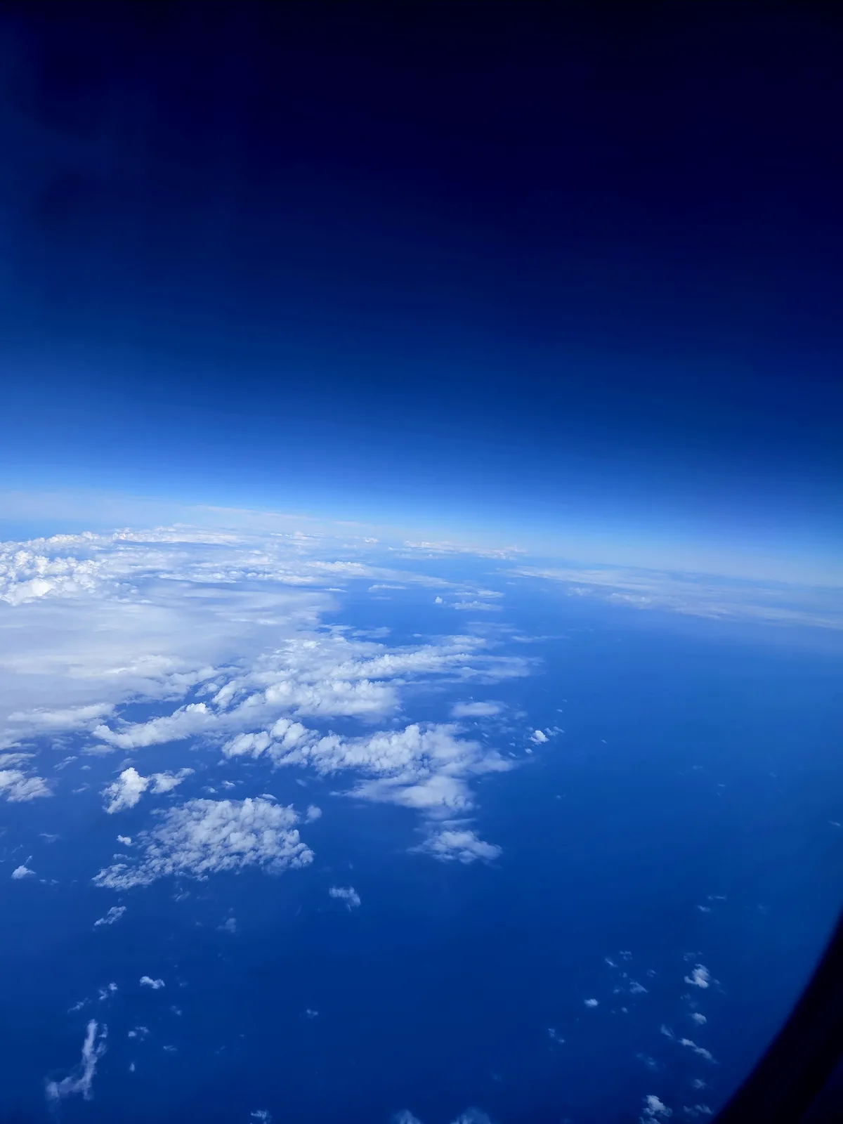 A view from an airplane window showing a vast expanse of deep blue sky and scattered white clouds below, with the curved…