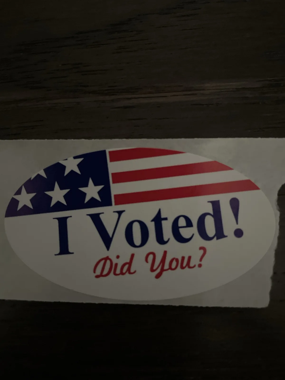 A close-up photo shows a standard oval-shaped 'I Voted!' sticker with an American flag design on a dark wooden surface. The…