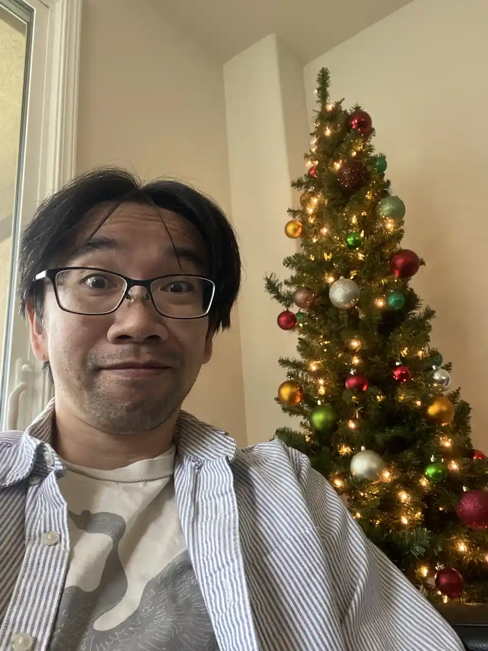A man with glasses and a striped shirt stares at the camera with a deadpan expression while standing in front of a decorated…