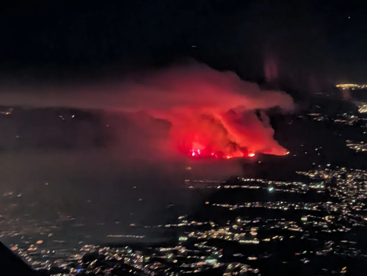 An aerial night view shows a massive, glowing red wildfire with thick smoke billowing over a sprawling cityscape illuminated…