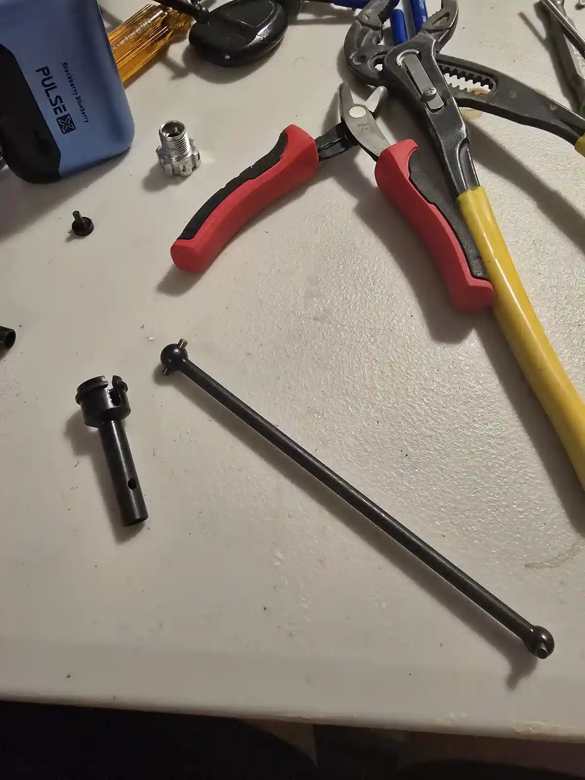 A cluttered workbench holds various tools including red-handled pliers, a yellow-handled wrench, and a black ball joint rod…