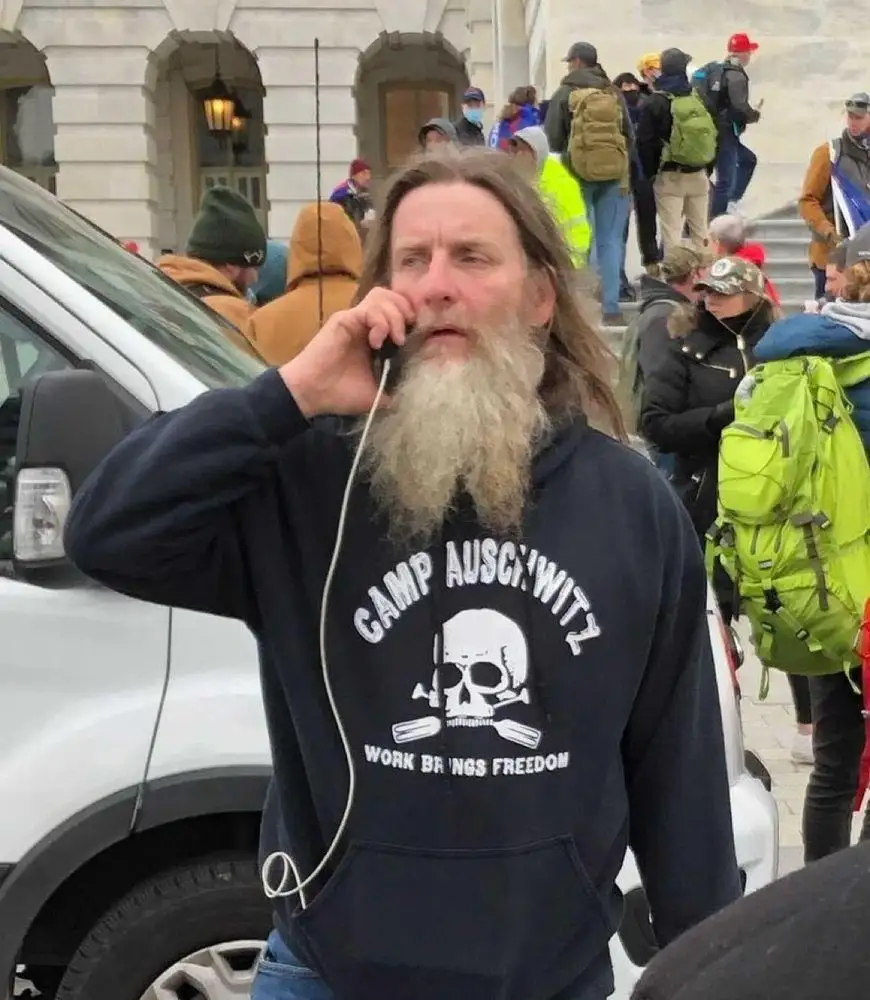 A man with a long beard and dark hoodie stands outdoors near a white vehicle, holding a phone to his ear. The hoodie…