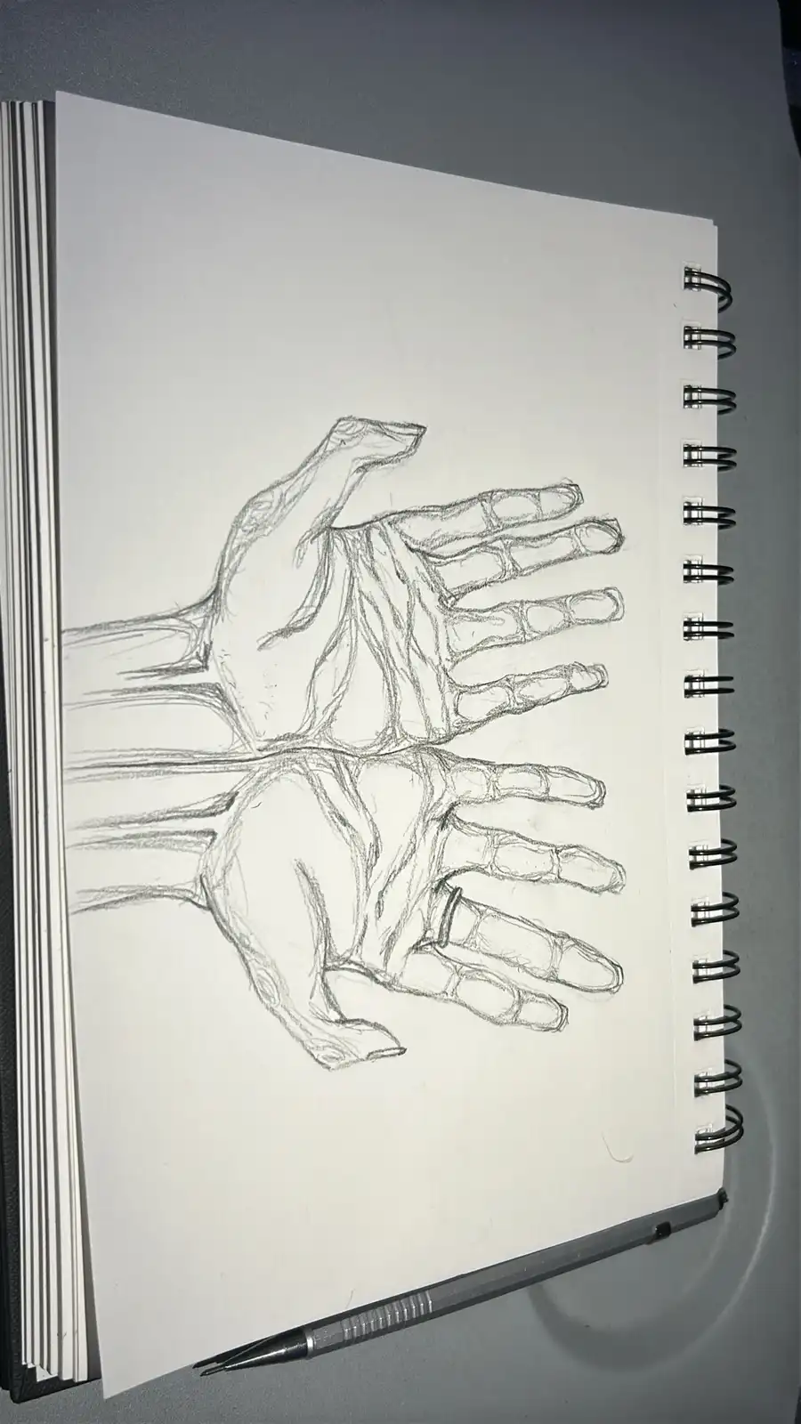 A pencil sketch in a spiral-bound sketchbook shows two hands drawn in a detailed, anatomical style with visible bones and…