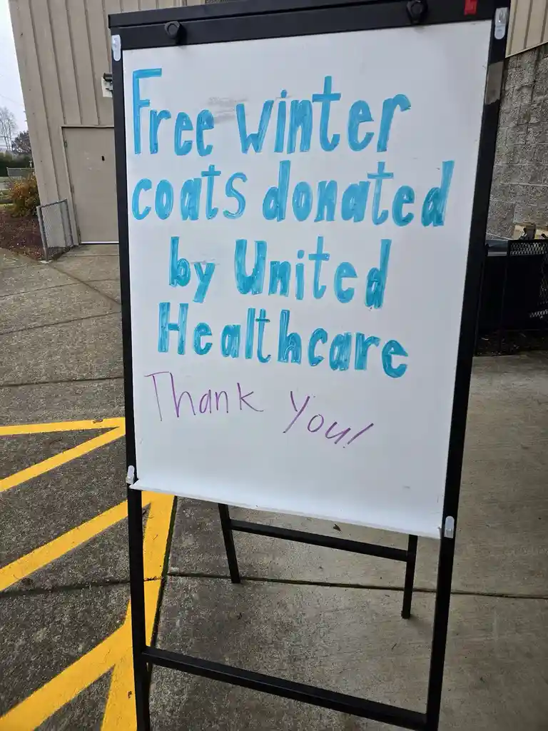 A whiteboard sign with blue and purple handwritten text stands outdoors on a sidewalk next to yellow parking lines. The sign…