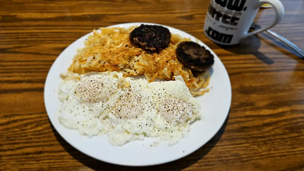 A plate of breakfast food on a wooden table, featuring fried eggs, crispy hash browns, and two burnt sausage patties. A…