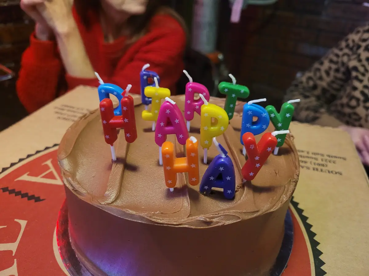 A chocolate birthday cake with colorful letter candles spelling 'HAPPY BIRTHDAY' sits on a cardboard box. In the blurred…