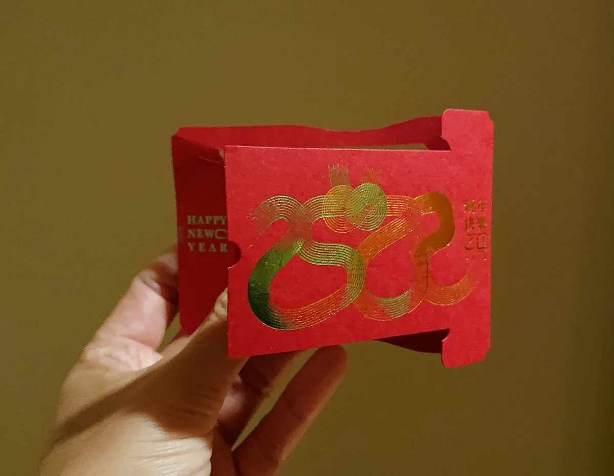 A hand holds a red, box-shaped Chinese New Year envelope with gold foil designs resembling a dragon or snake, and the text…