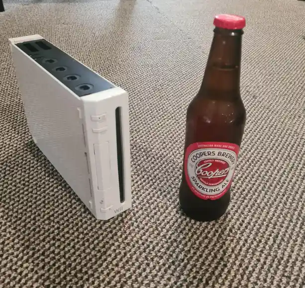 A white Nintendo Wii console sits on a patterned carpet next to a brown Coopers Sparkling Ale beer bottle with a red cap and…