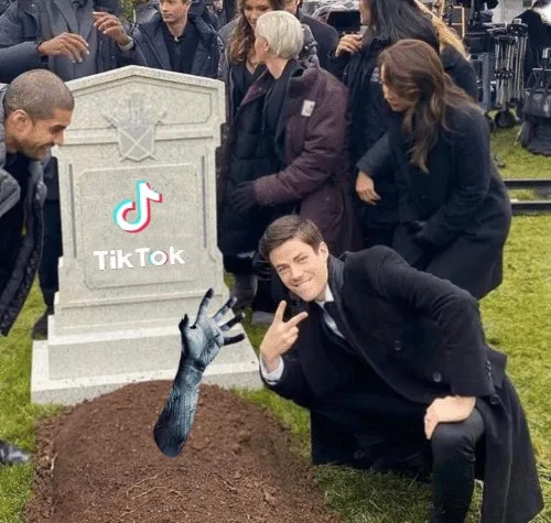 A man in a suit is kneeling beside a freshly dug grave, smiling and making a peace sign with one hand while a gloved hand…
