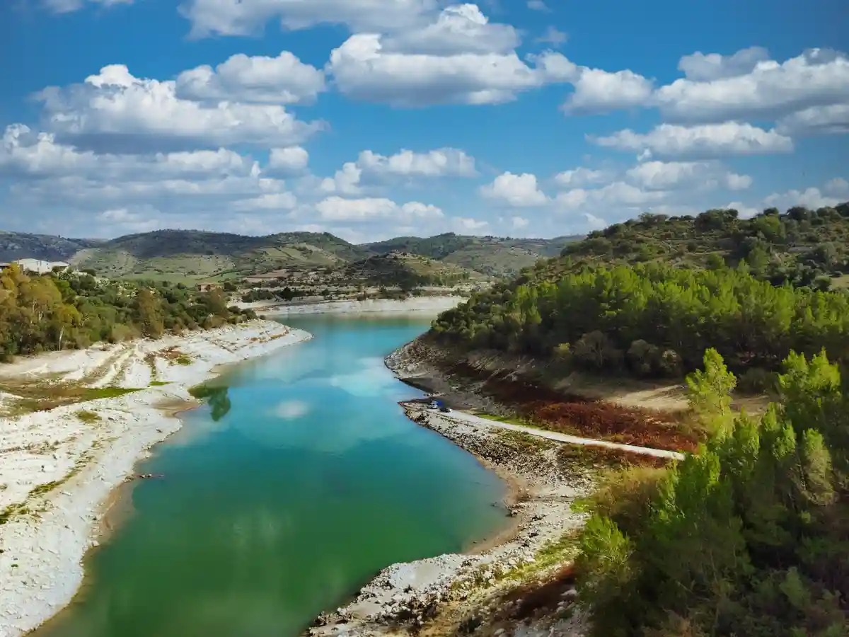An aerial view of a narrow, turquoise lake surrounded by green hills and trees under a blue sky with white clouds. The water…