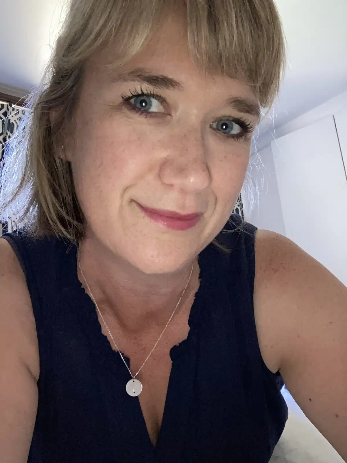 A selfie of a woman with blonde hair and blue eyes, wearing a dark blue sleeveless top and a silver necklace with a circular…