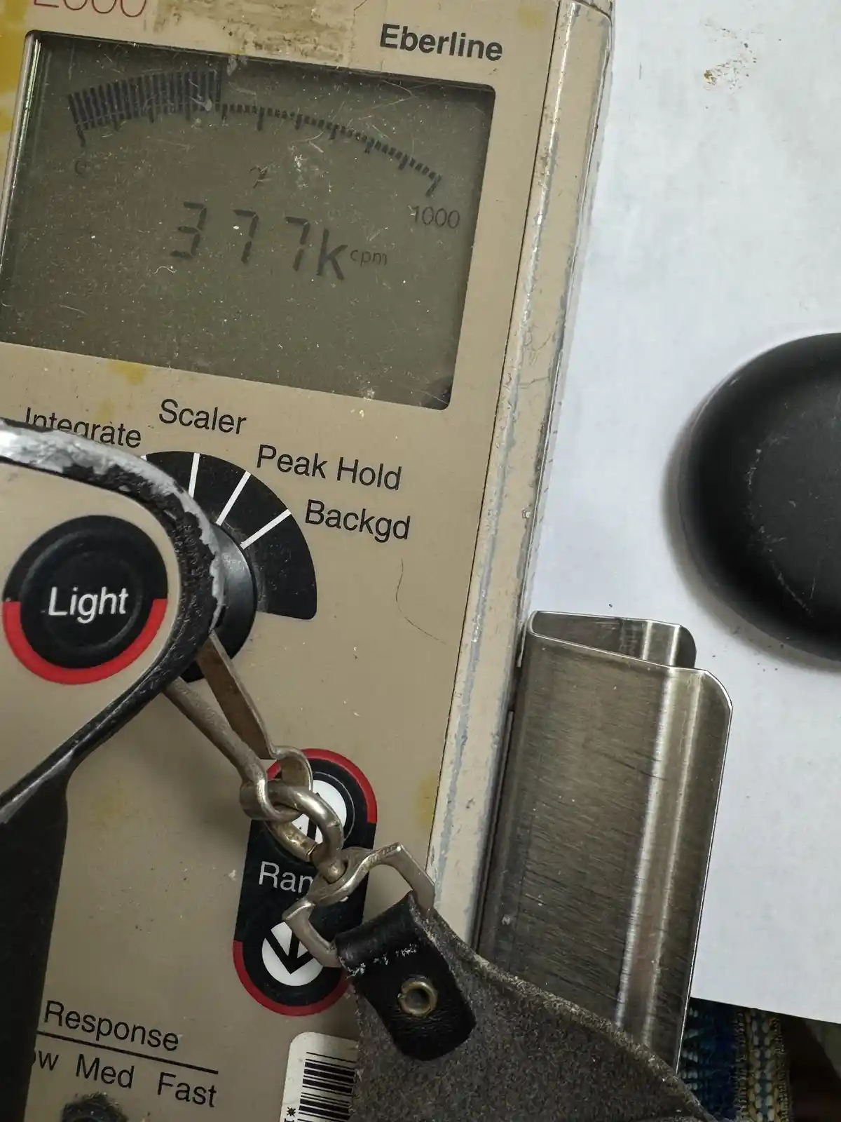 A close-up of an Eberline radiation detector showing a reading of 377k cpm on its digital display, with a metal tool hanging…