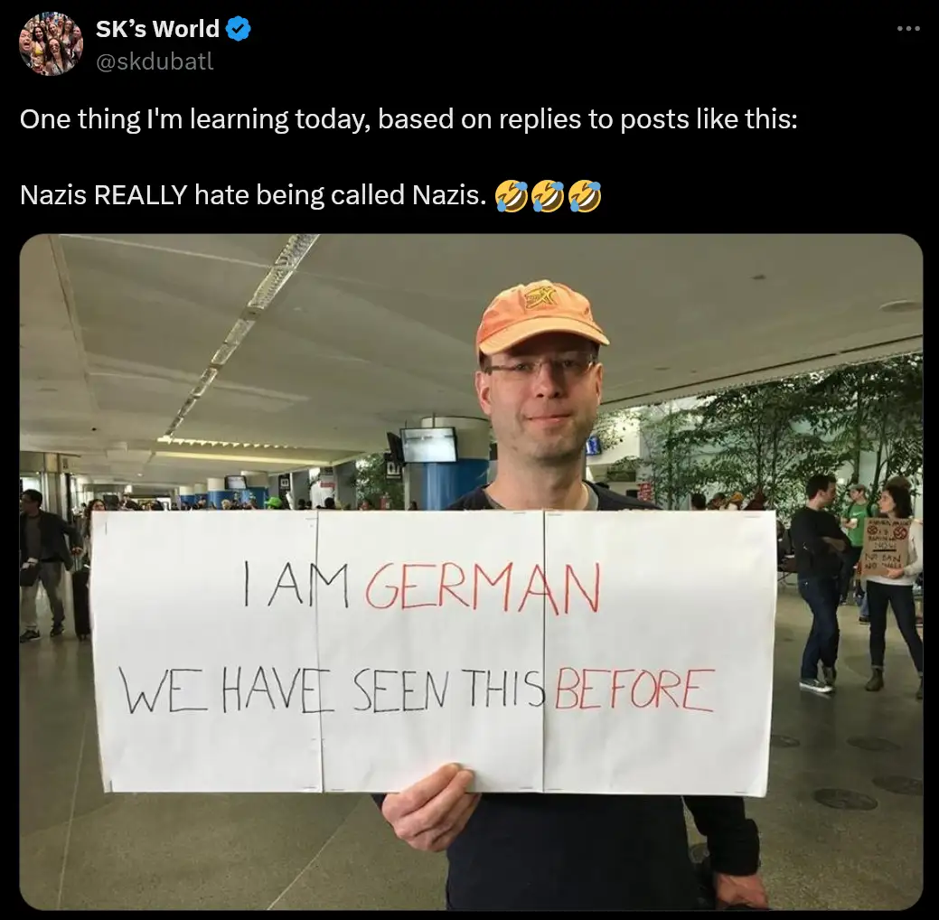A man in an orange cap and glasses holds a large white sign in a public indoor space, likely an airport. The sign has…