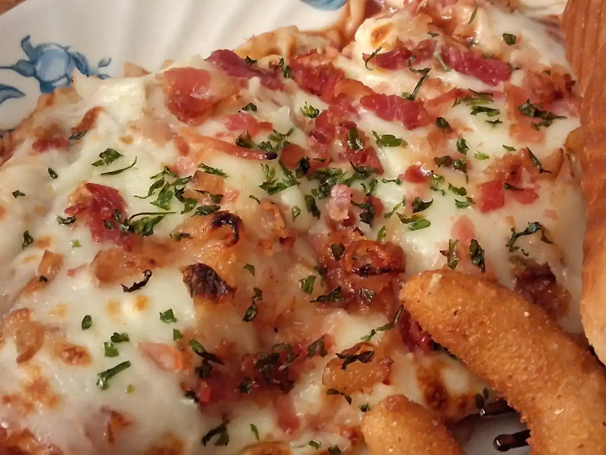 A close-up photo of a cheesy dish topped with bacon bits, green herbs, and crispy fried food items, served on a plate with a…