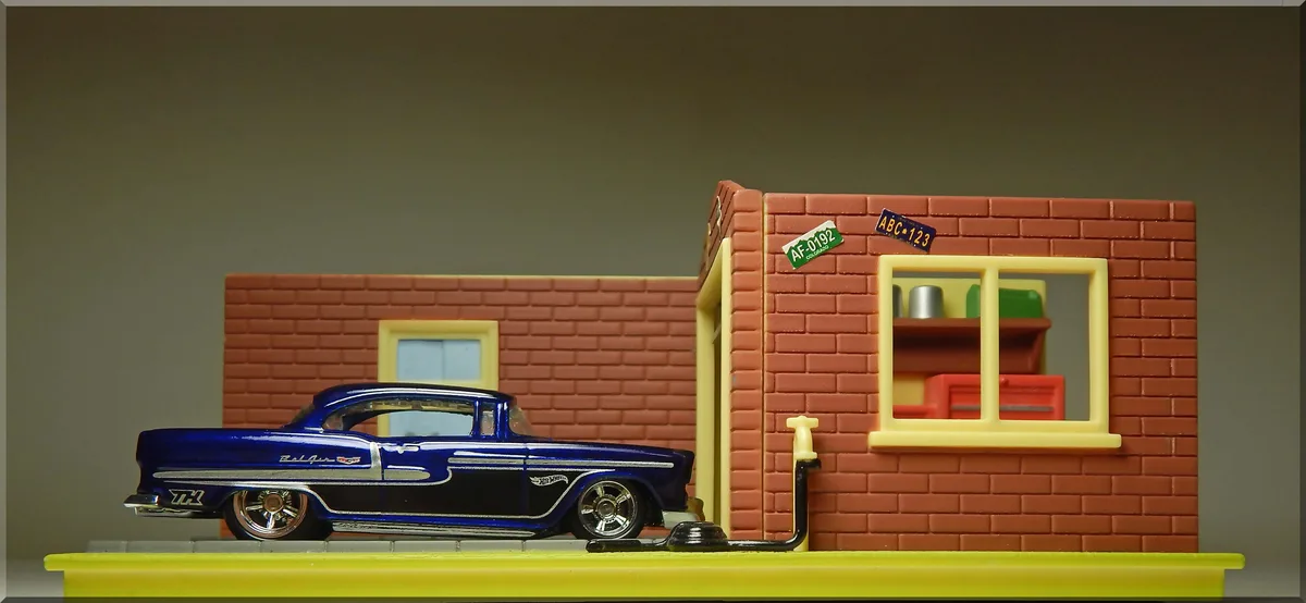 A blue Hot Wheels model car is parked beside a small brick building with a yellow window frame and a black hose extending…
