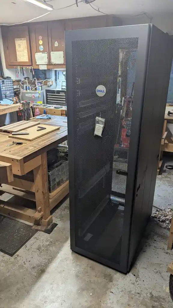 A large black Dell server cabinet with a mesh front door stands in a cluttered home workshop filled with wooden workbenches…