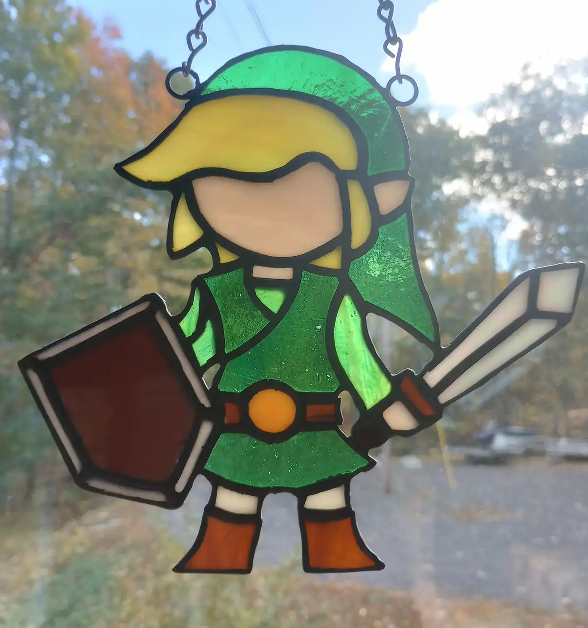 A stained glass window hanging depicts a chibi-style Link from The Legend of Zelda, wearing green tunic and hat, holding a…