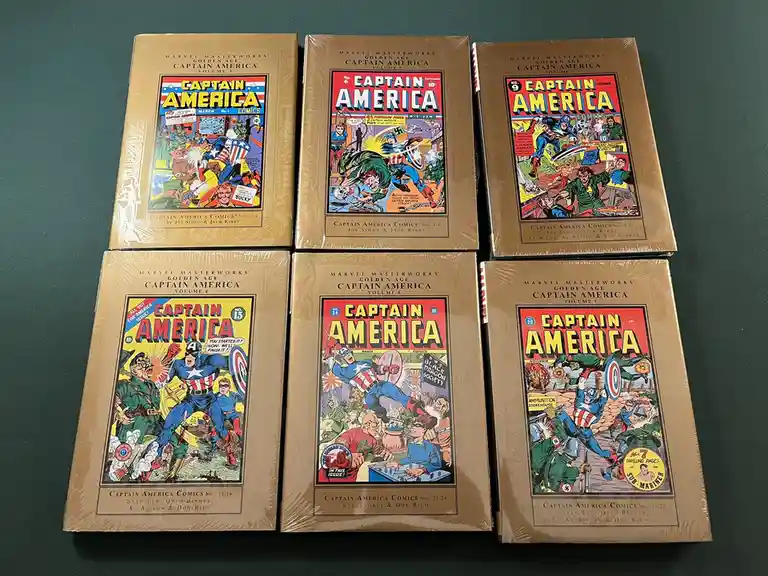 Six sealed Marvel Masterworks Golden Age Captain America comic book volumes are arranged in two rows on a dark green…