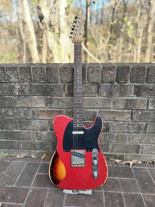 A red and black Fender Telecaster electric guitar with significant wear and tear, including a large hole in the body, is…
