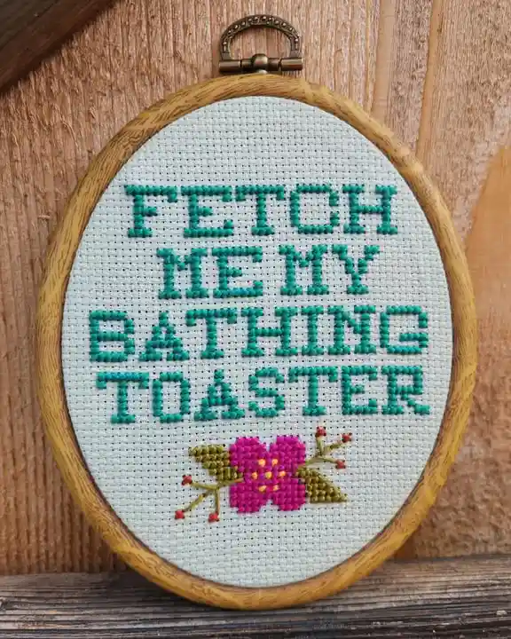 A wooden embroidery hoop holds a piece of white fabric with teal cross-stitch text reading 'FETCH ME MY BATHING TOASTER' and…