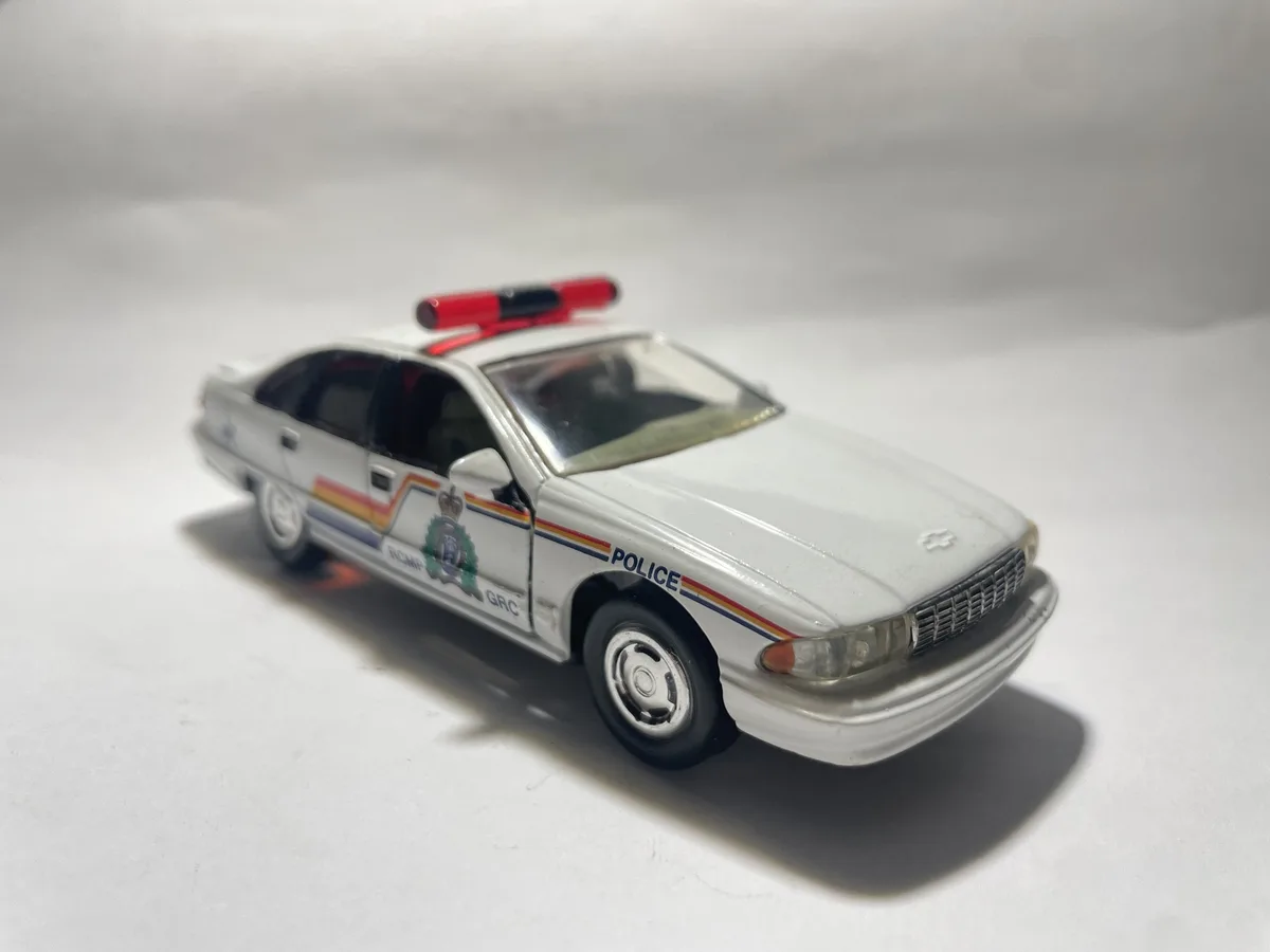 A white toy police car with 'POLICE' and 'RCMP GRC' markings, a red light bar, and rainbow stripes is photographed on a…