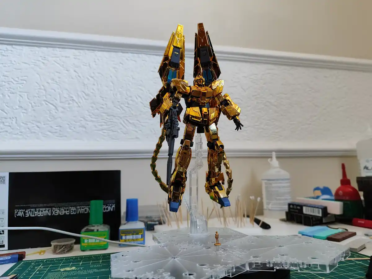 A highly detailed, gold-painted Gundam model stands on a clear plastic display stand on a cluttered hobbyist workbench. The…