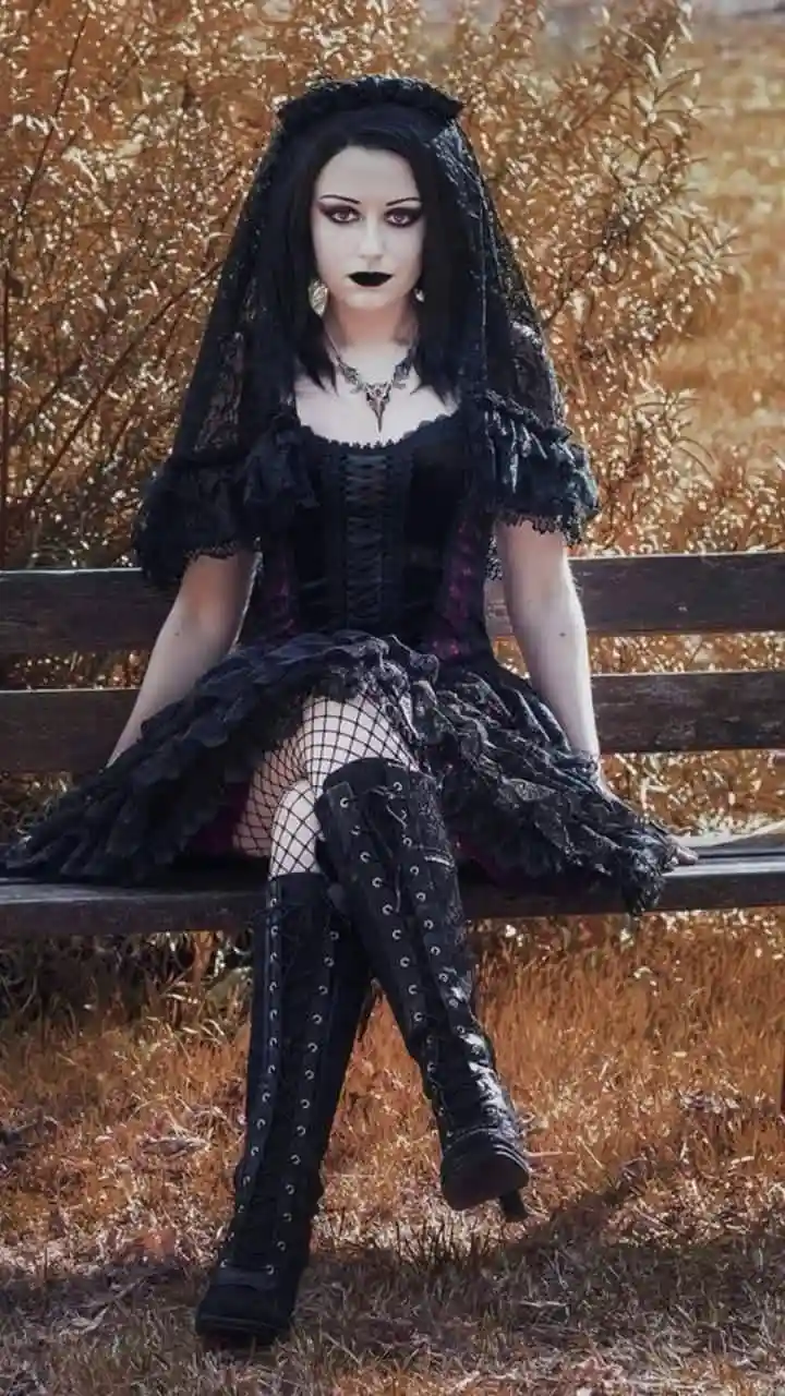 A woman dressed in full gothic attire — black lace veil, corset dress, fishnet stockings, and tall boots — sits on a park…